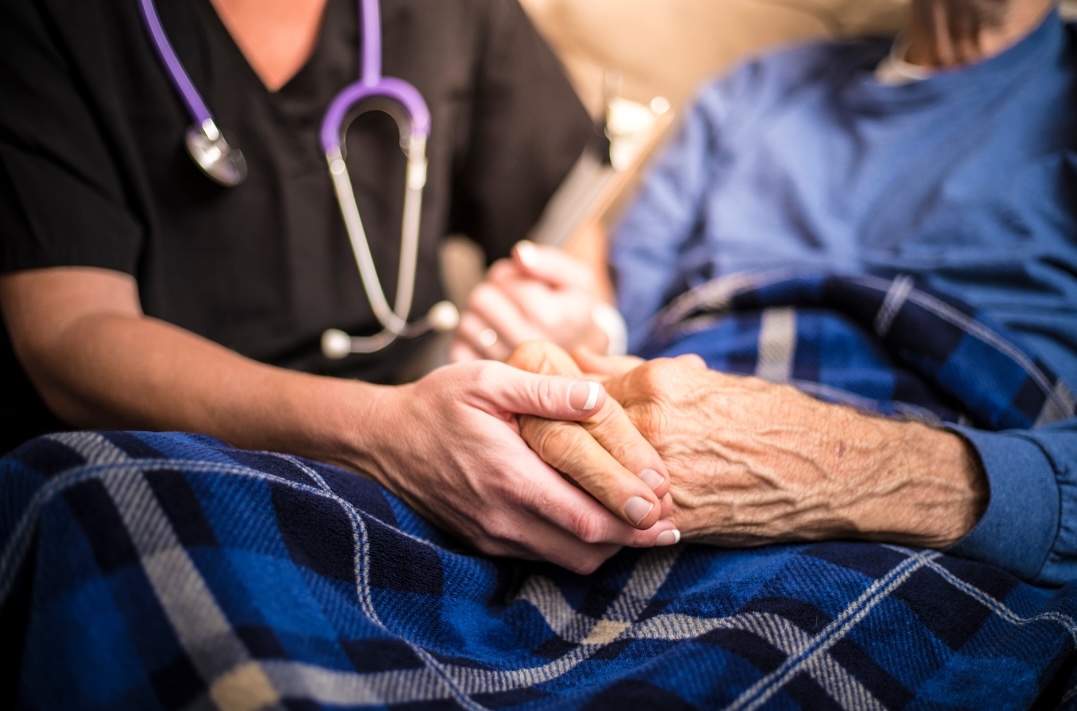 A medical worker, perhaps a hospice worker or nurse, holds hands with an elderly man. Her stethoscope appears next to his blue pajamas.